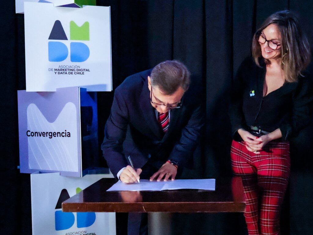 Man in a suit signs a document at a podium while a woman in red plaid pants smiles beside him at a formal event with marketing signage in the background.