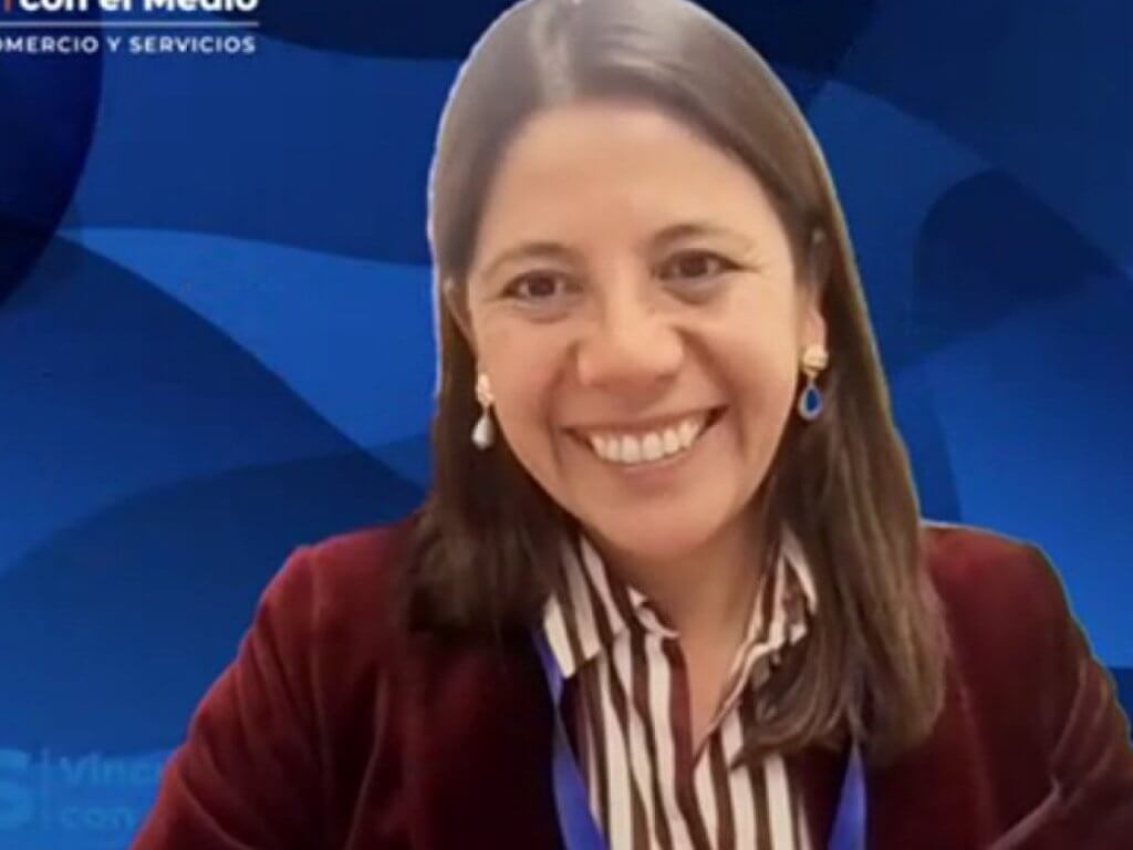 Smiling woman in a maroon blazer and striped blouse on a video broadcast with a blue geometric studio backdrop.