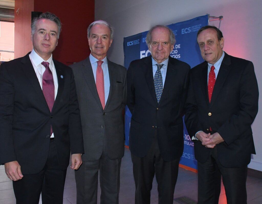 Four men in business suits posing together at a formal event indoors, with a blue banner in the background.
