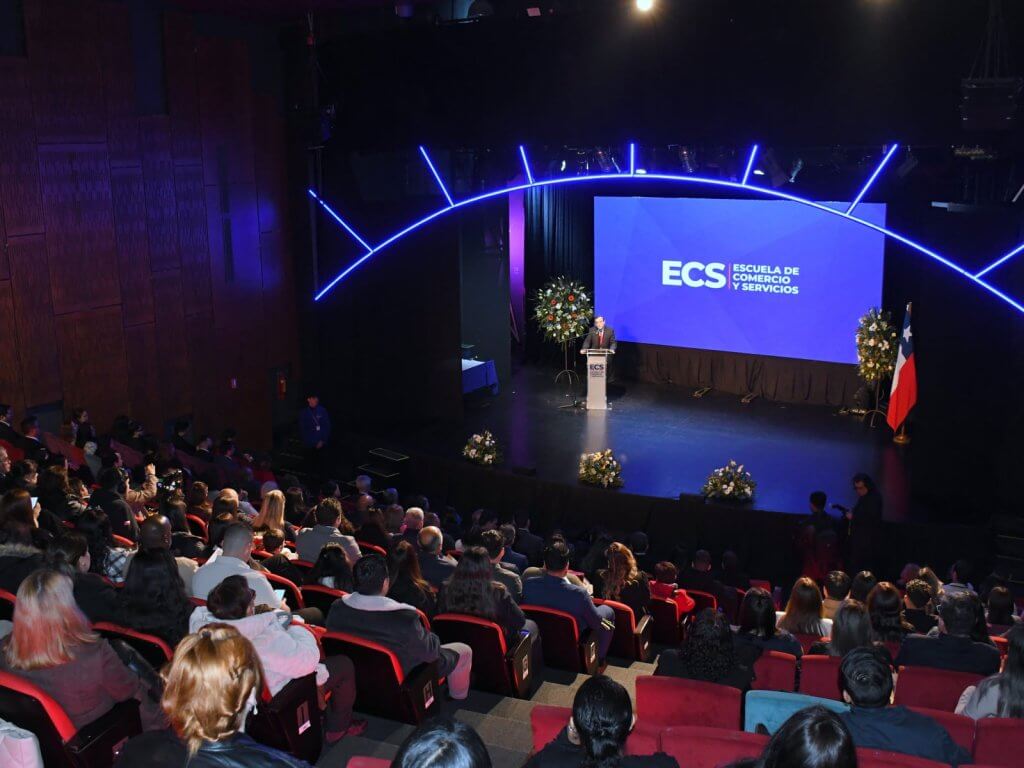 Speaker at podium on stage delivering presentation to a large audience in an auditorium with a blue ECS screen and Chile flag nearby.