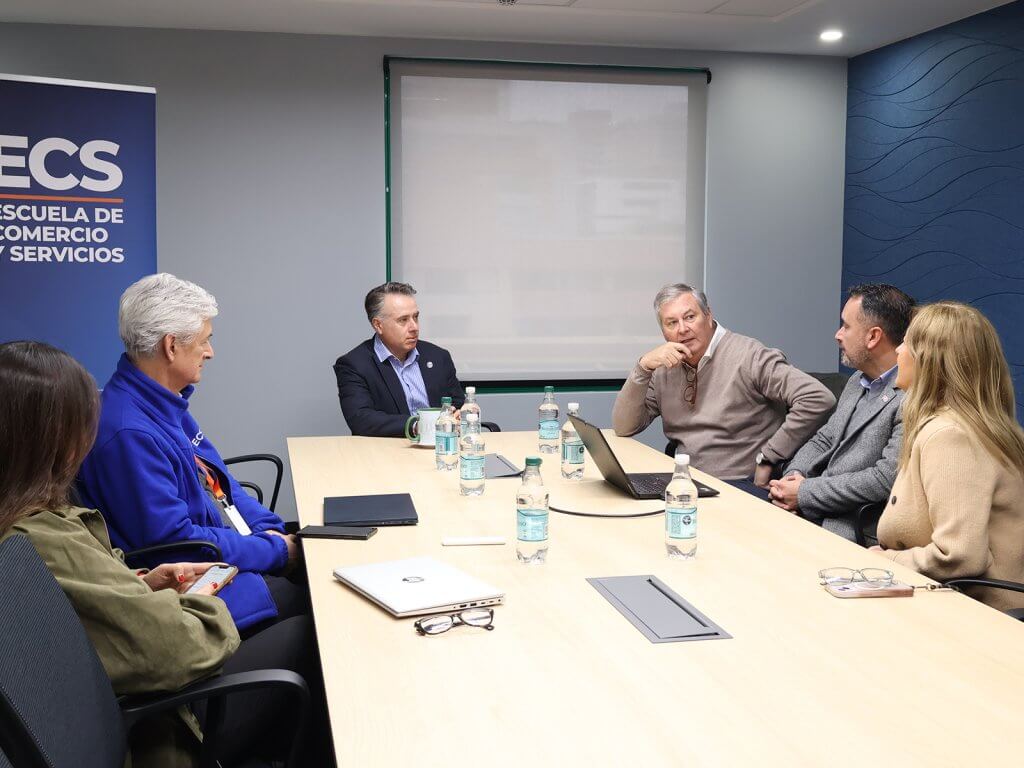 Six people in a business meeting around a conference table; water bottles, laptops, and a blue banner on the left reading Escuela de Comercio (ECS).** This is incorrect due to formatting; adjust to clean text