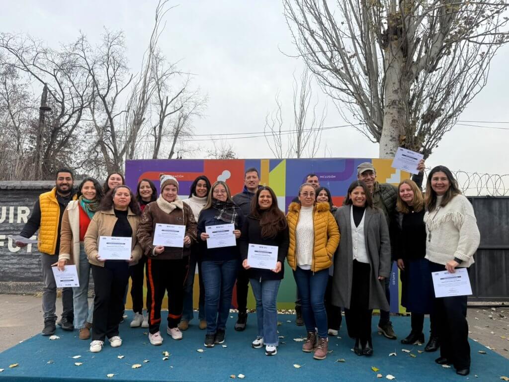 Group of people holding certificates on a blue stage outdoors, smiling after a ceremony, colorful mural in the background.