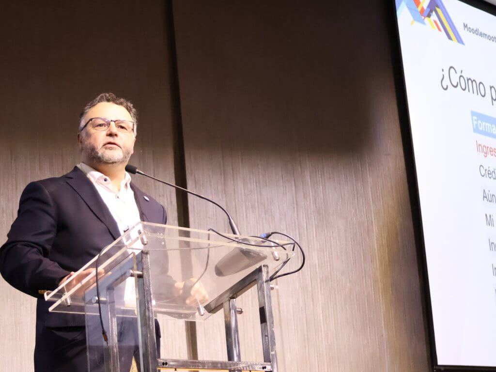 Male presenter in a dark blazer speaks at a clear lectern with a microphone, slides shown on a large screen to the right.