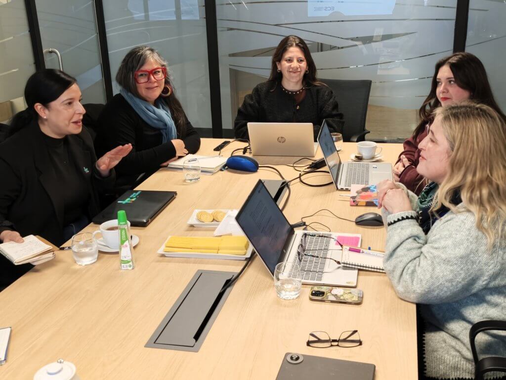 Five women sit around a conference table in a glass-walled meeting room, laptops open and notes in hand during a discussion.