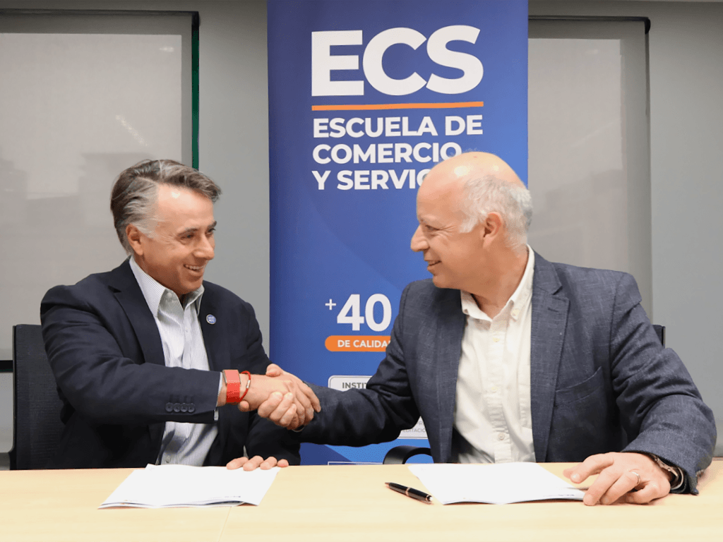 Two men in business suits shake hands over documents on a conference table with a blue banner behind them that reads Escuela de Comercio y Servicios.