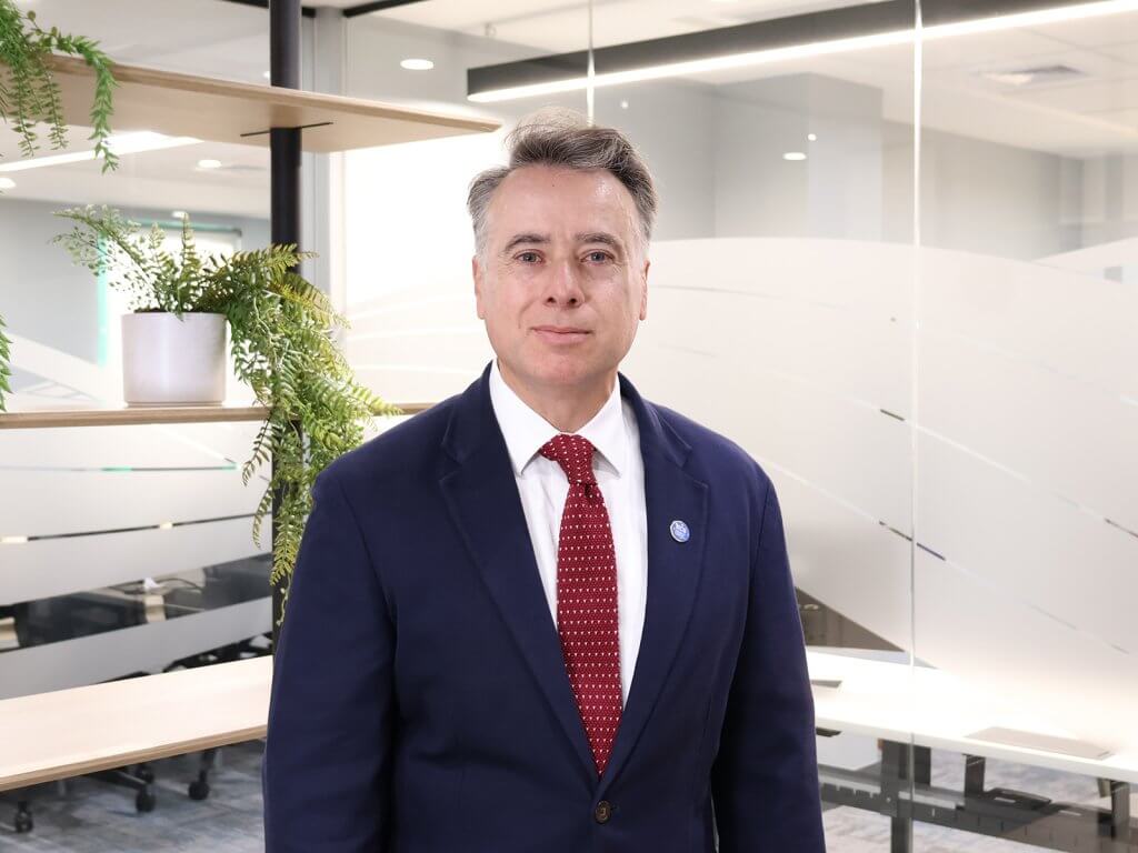 Portrait of a man in a navy suit, white shirt, and red tie standing in a bright office with glass walls and plants behind him.