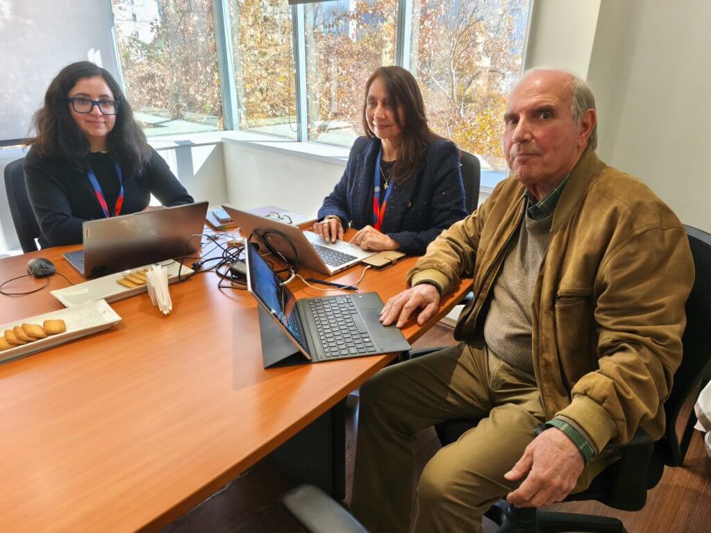 Three colleagues sit around a wooden conference table with laptops, snacks, and cables in a bright office, autumn trees outside the window.