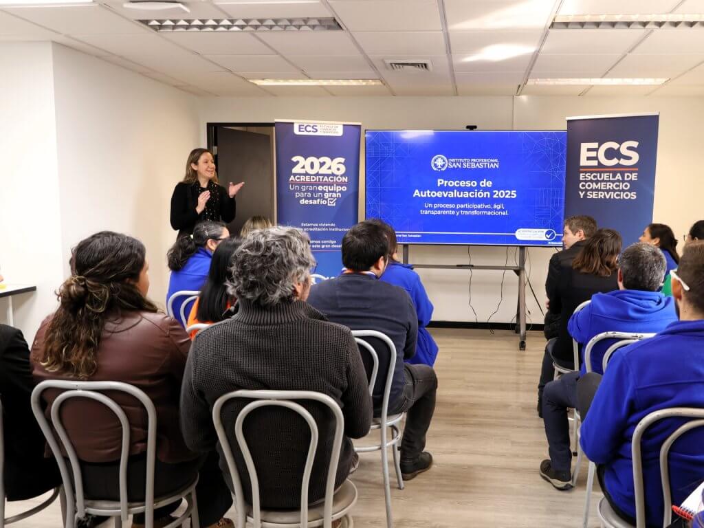 Speaker presenting to an audience in a classroom-style event with blue banners and a large screen displaying a slide about evaluation process and accreditation](alt2)