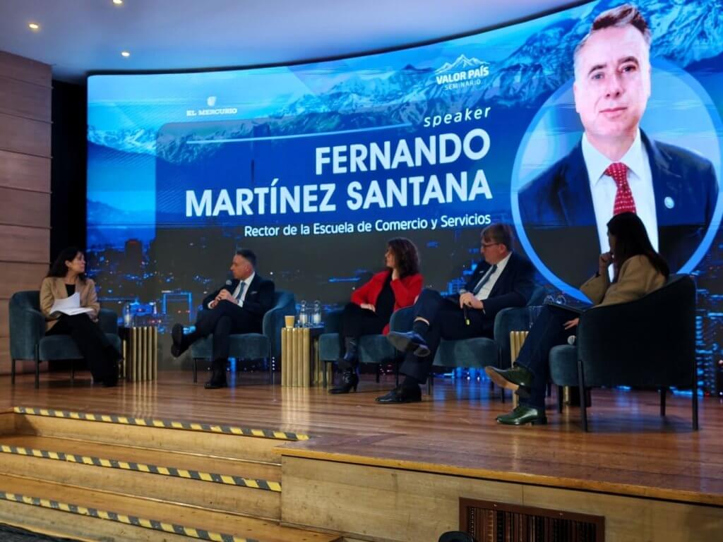 Panel of five speakers on a stage with a large blue banner backdrop featuring the name 'Fernando Martínez Santana' and a portrait on the right side. Conducting a moderated discussion at a conference event.