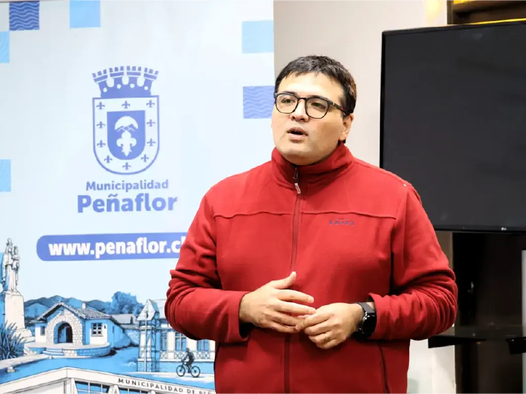 Male presenter in a red jacket speaks to an audience in front of a Municipalidad de Peñaflor backdrop.