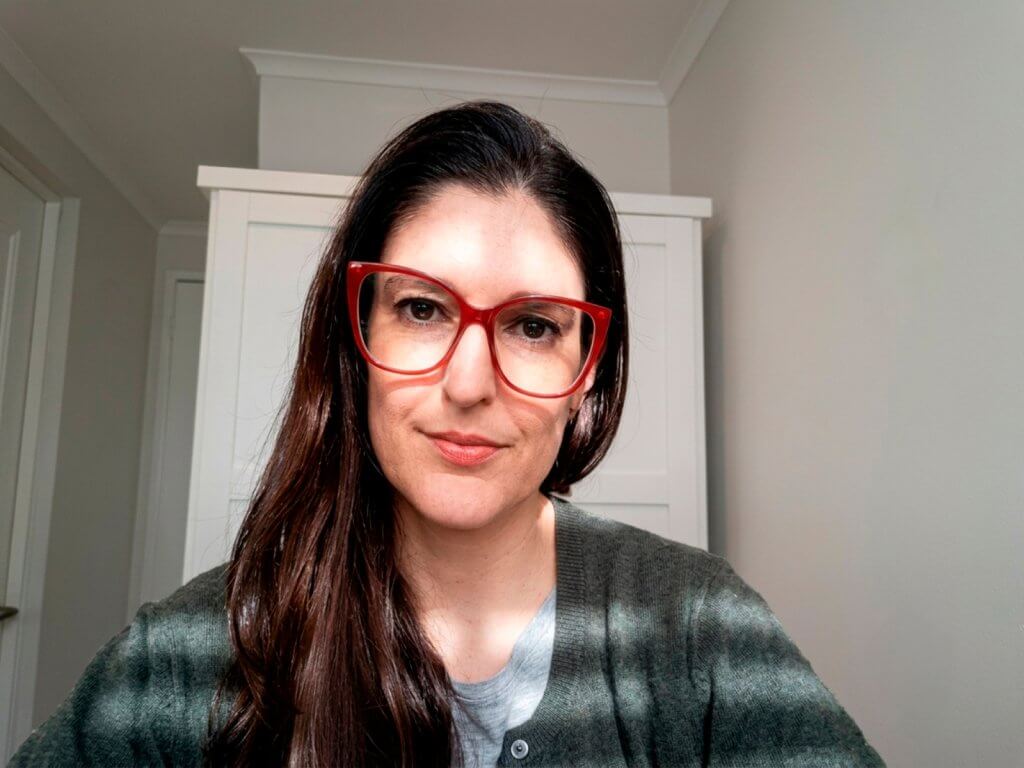 Self-portrait of a woman with red glasses, wearing a gray-green cardigan, indoors with a white cabinet in the background.