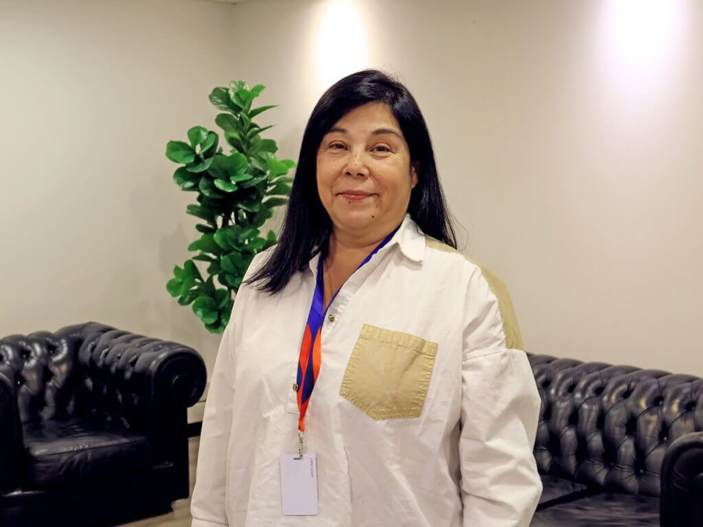 Smiling woman in a white shirt with a beige pocket, standing in a lounge with a green plant behind her.