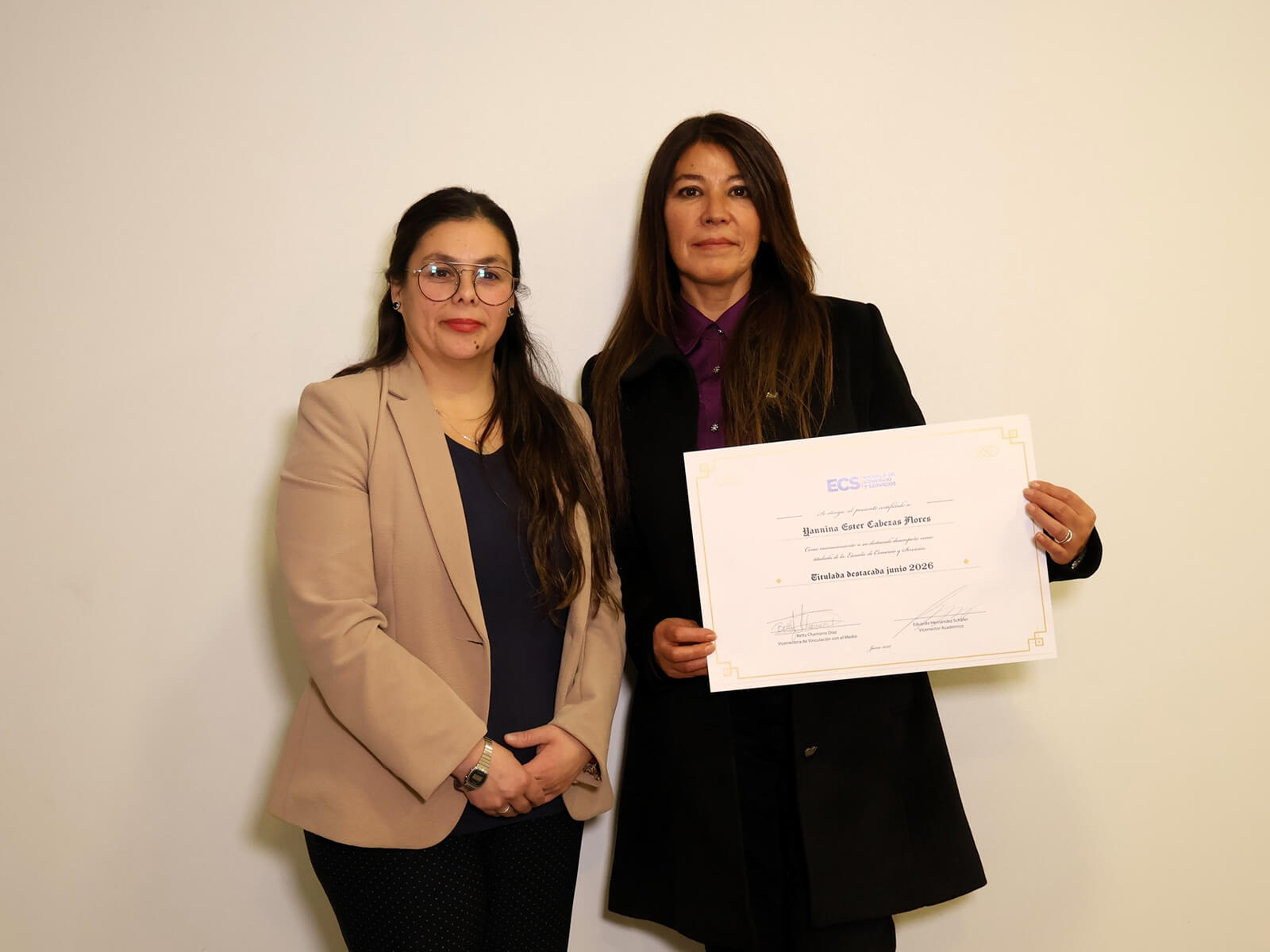 Two women pose against a light wall; one holds a large certificate with a gold border.