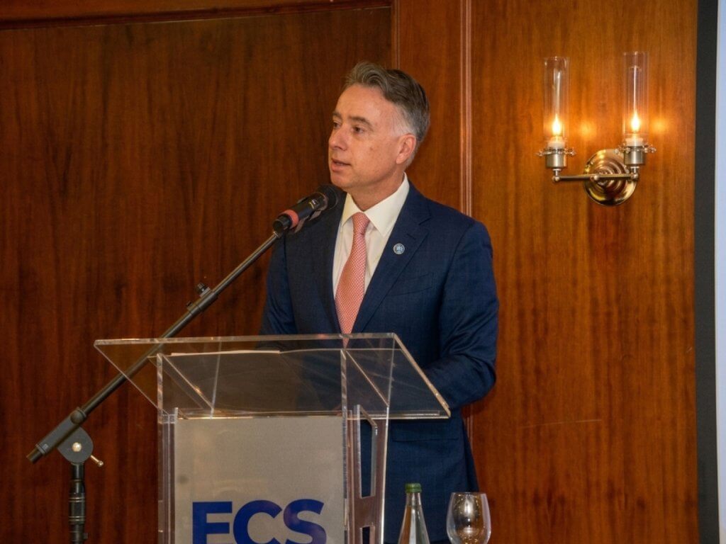 Man in navy suit and pink tie speaks at a clear acrylic podium in a wood-paneled room, ECS signage visible on the front.